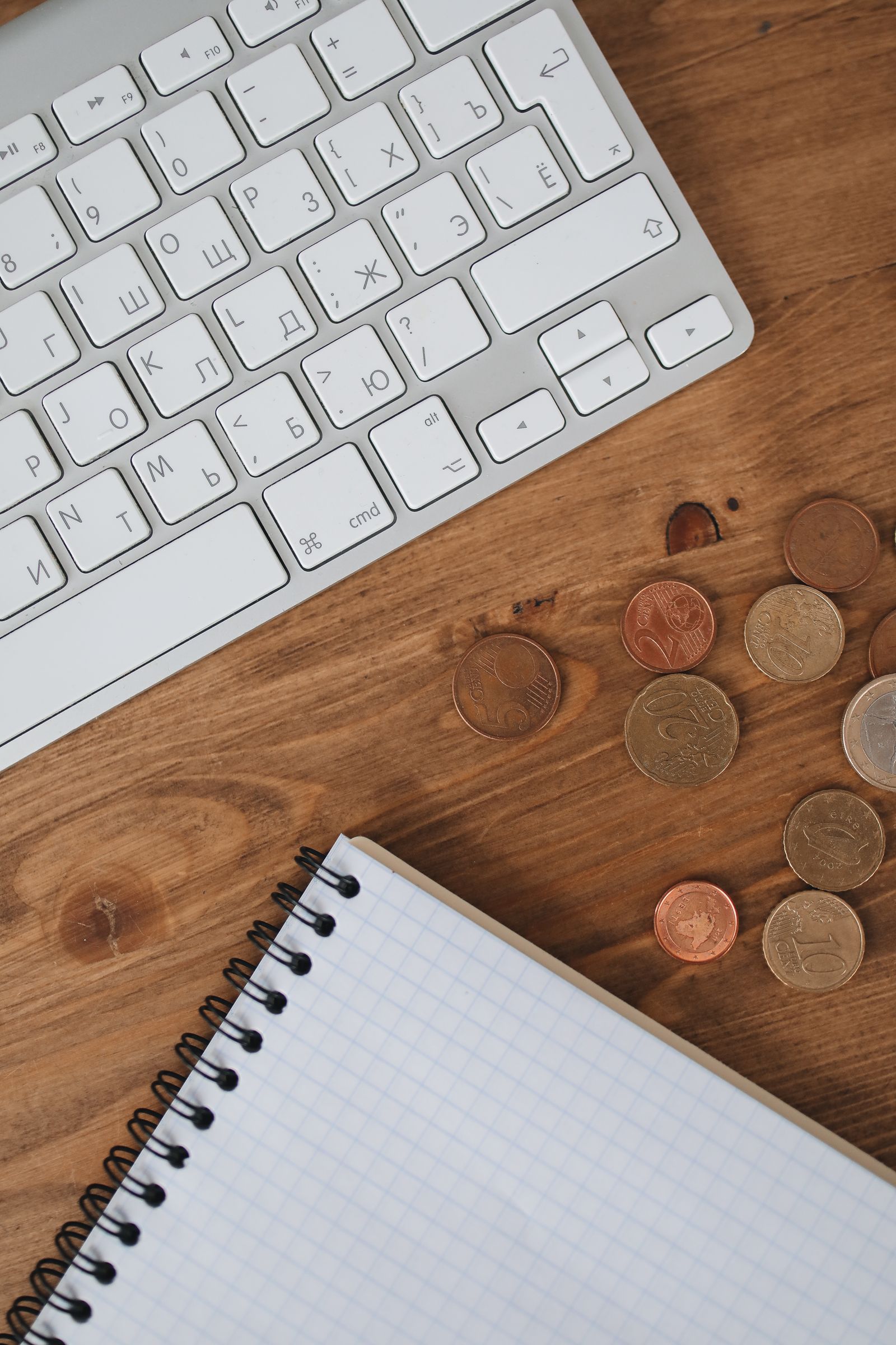 A keyboard, a grid notebook, and a small pile of coins on a wooden desk.