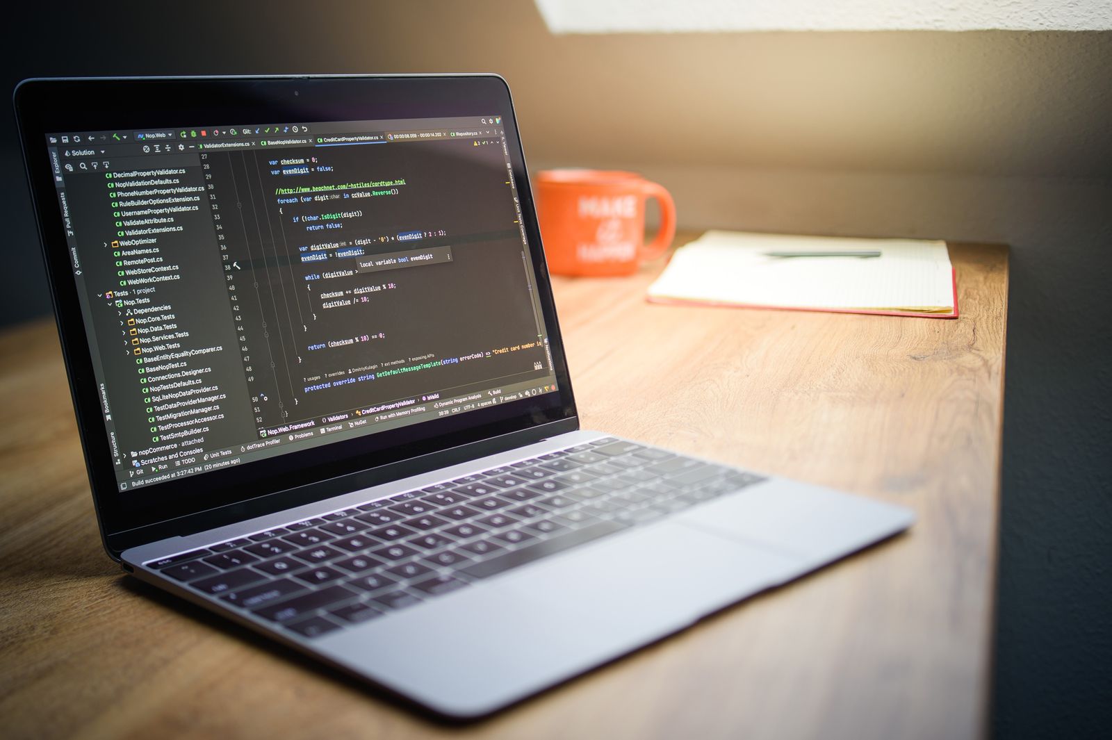 Laptop on a wooden desk showing a code editor, next to an orange mug and a notebook