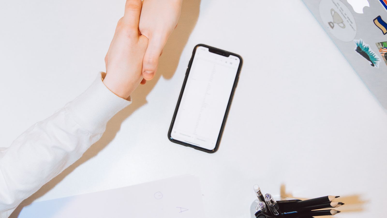 A handshake over a clean white desk with a phone and pens, representing client onboarding