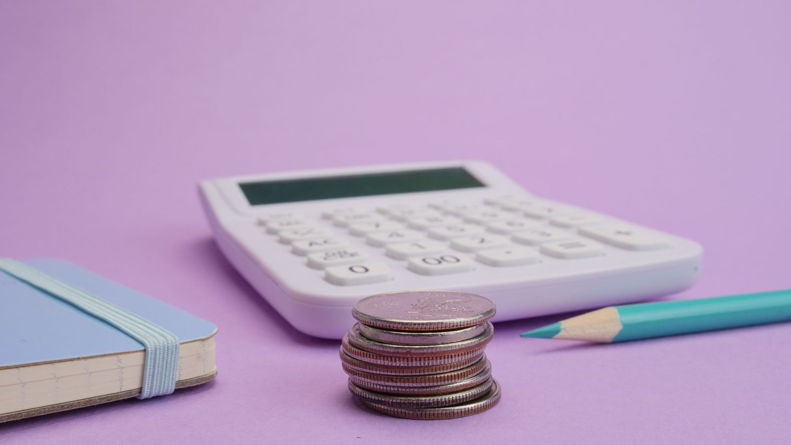 A white calculator, a stack of coins and a notebook on a purple background