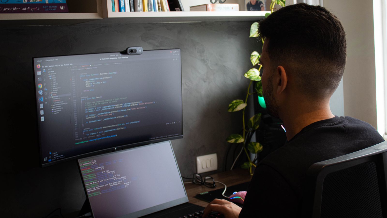 Developer at a home desk with a code editor and terminal open on a large monitor and laptop
