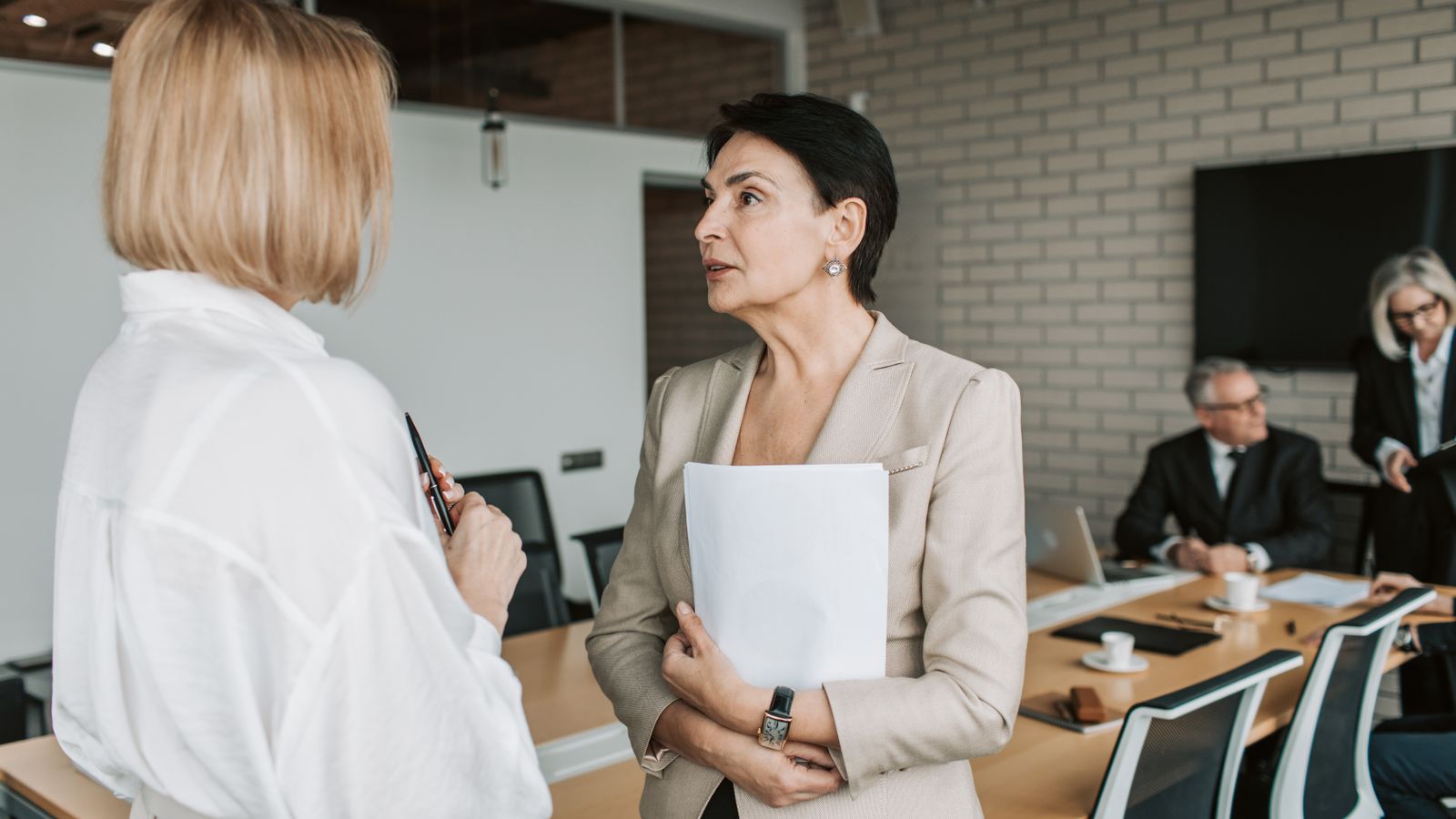 Two people talking in a meeting room with colleagues at a table behind them.