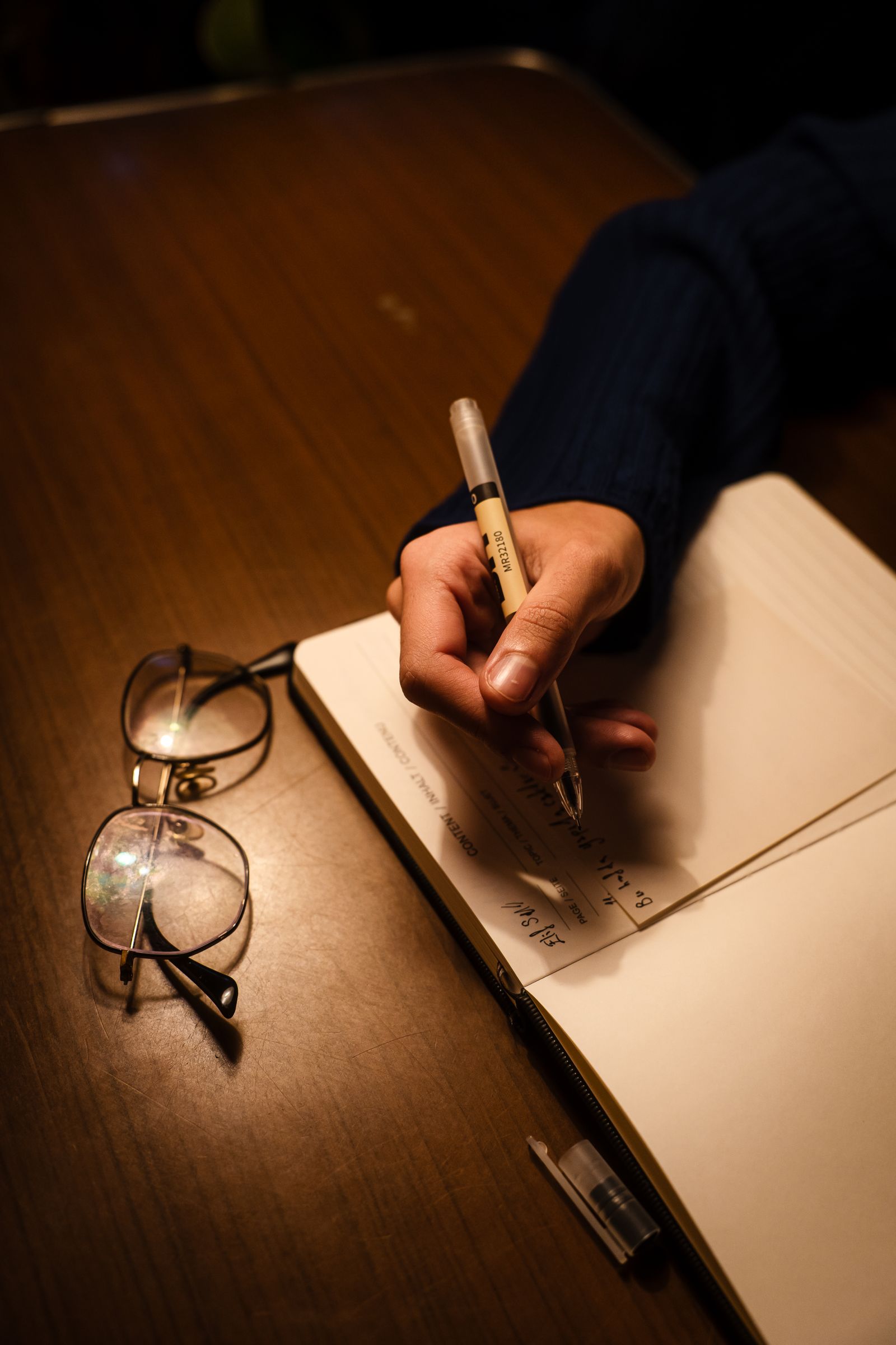 Person writing notes in a hardcover journal with reading glasses resting beside the page